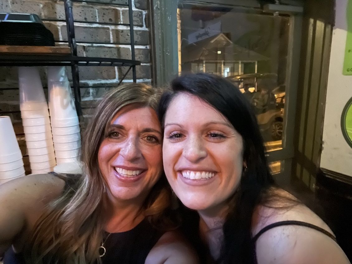 Two smiling women taking a selfie indoors, with a brick wall and outdoor view visible.
