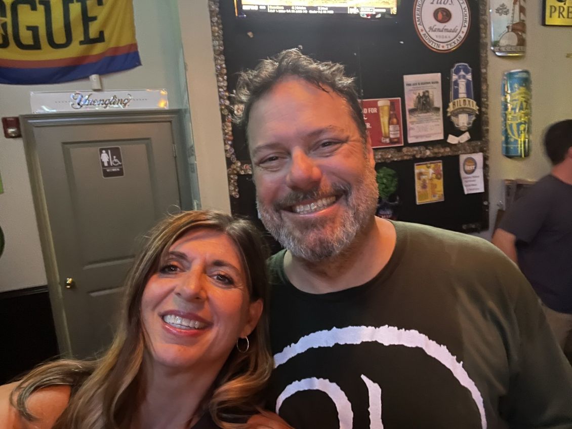 Smiling couple posing for a selfie inside a bar. Woman with long hair and man with beard. Background with bar decor.