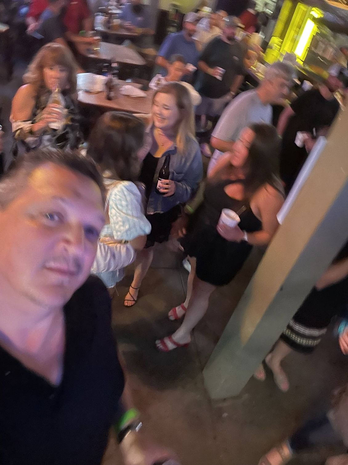 A crowded bar scene; people socializing, holding drinks. Man takes a selfie in the foreground.