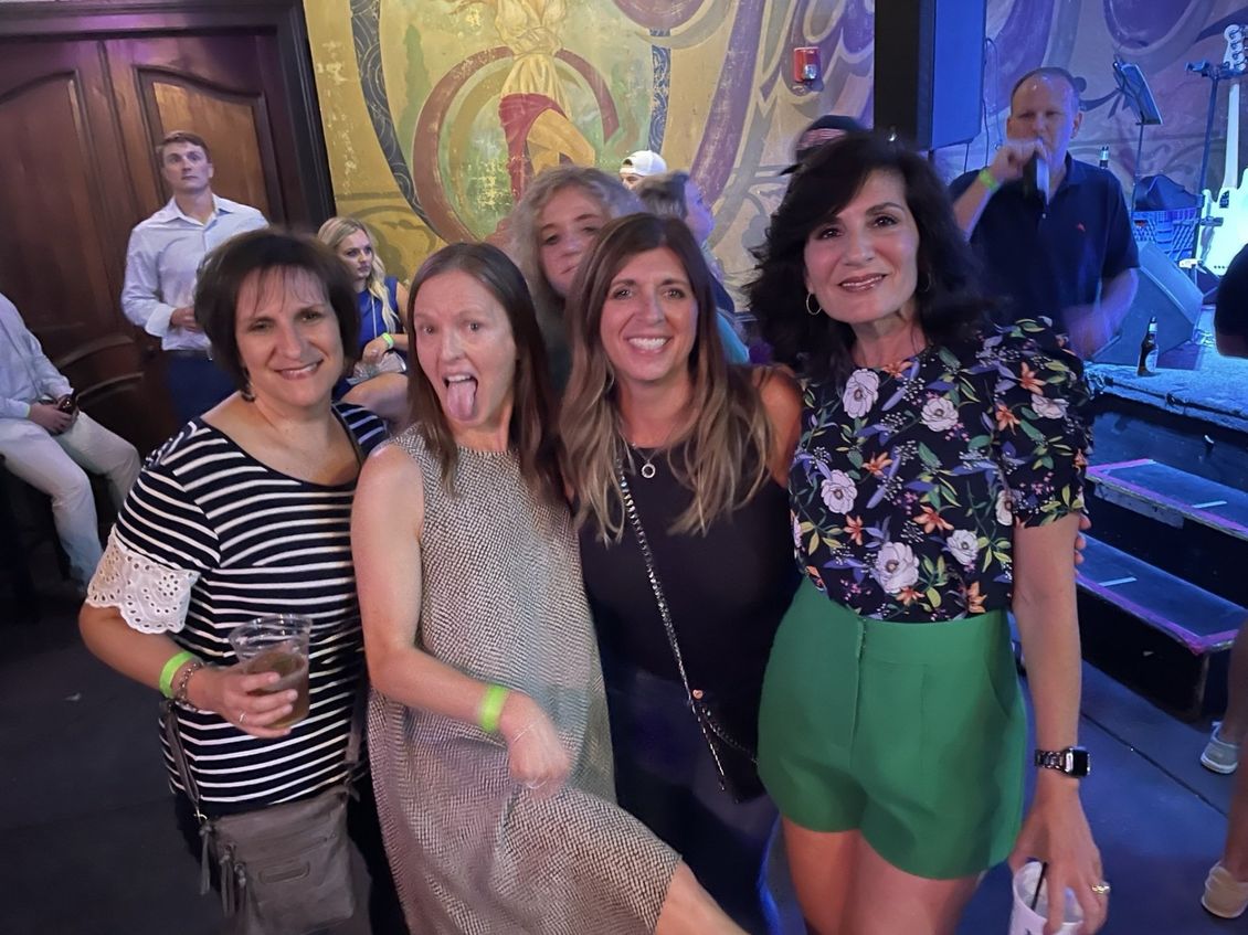 Group of women posing together in a bar, one sticking out her tongue, others smiling, mural in the background.