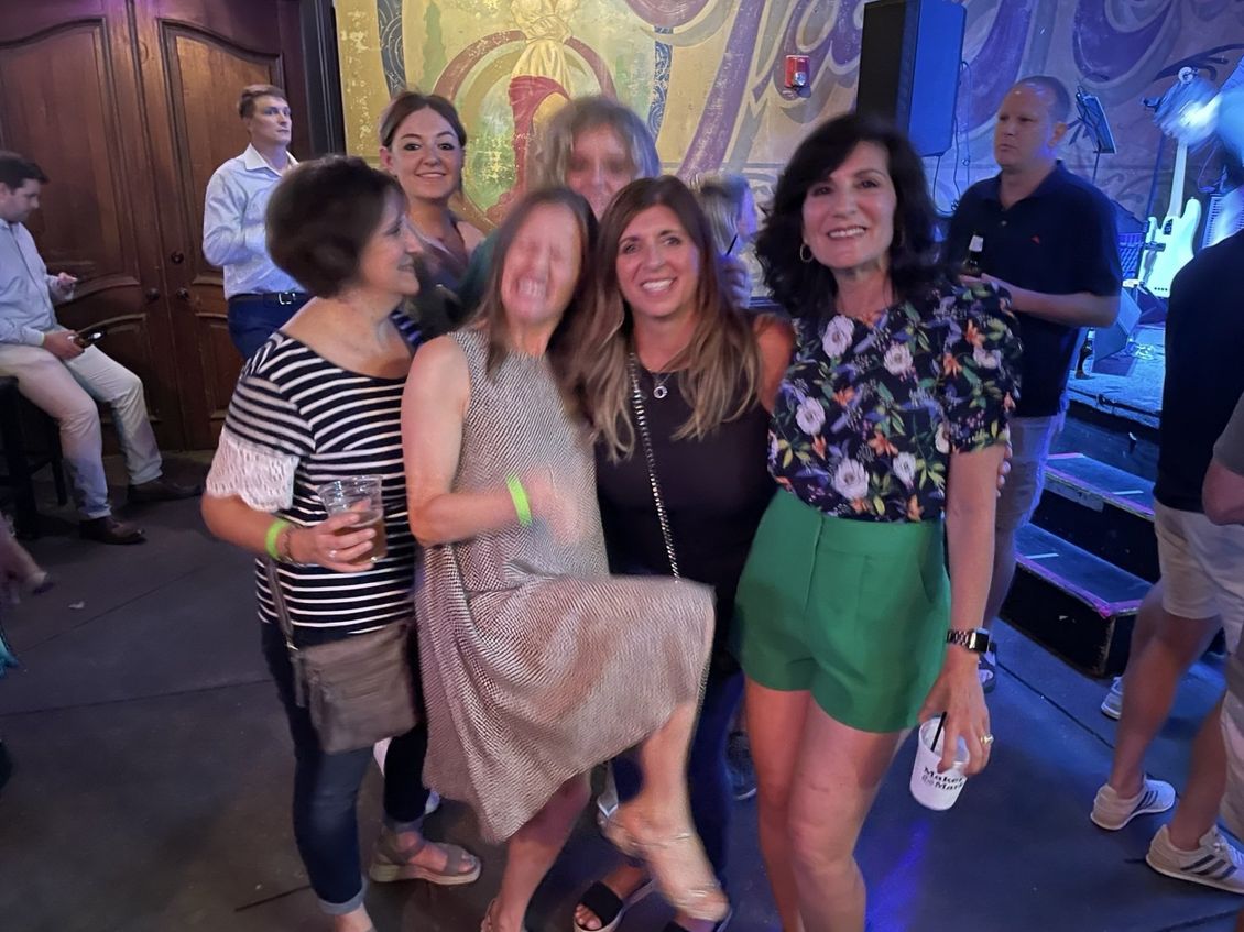 Group of smiling people at an event, posing with drinks. One woman playfully kicks up a leg.
