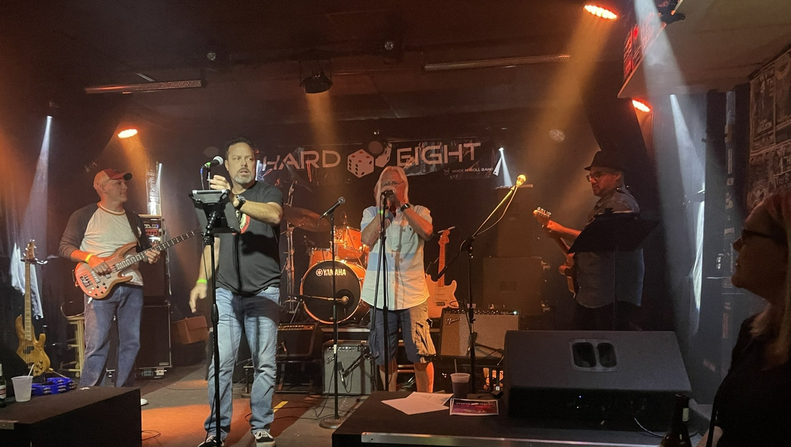 Band performing on stage with microphones and instruments. Low lighting, orange/yellow hues, small venue.