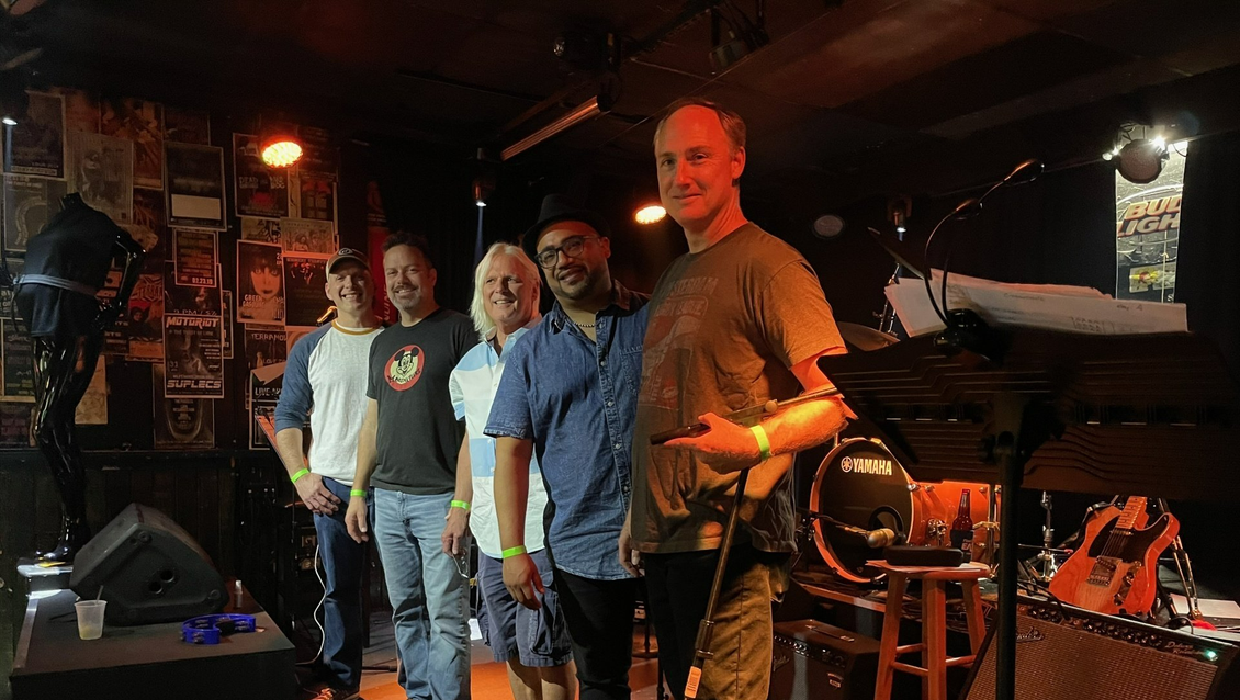 Band of five men standing onstage at a bar, lit by orange stage lights.