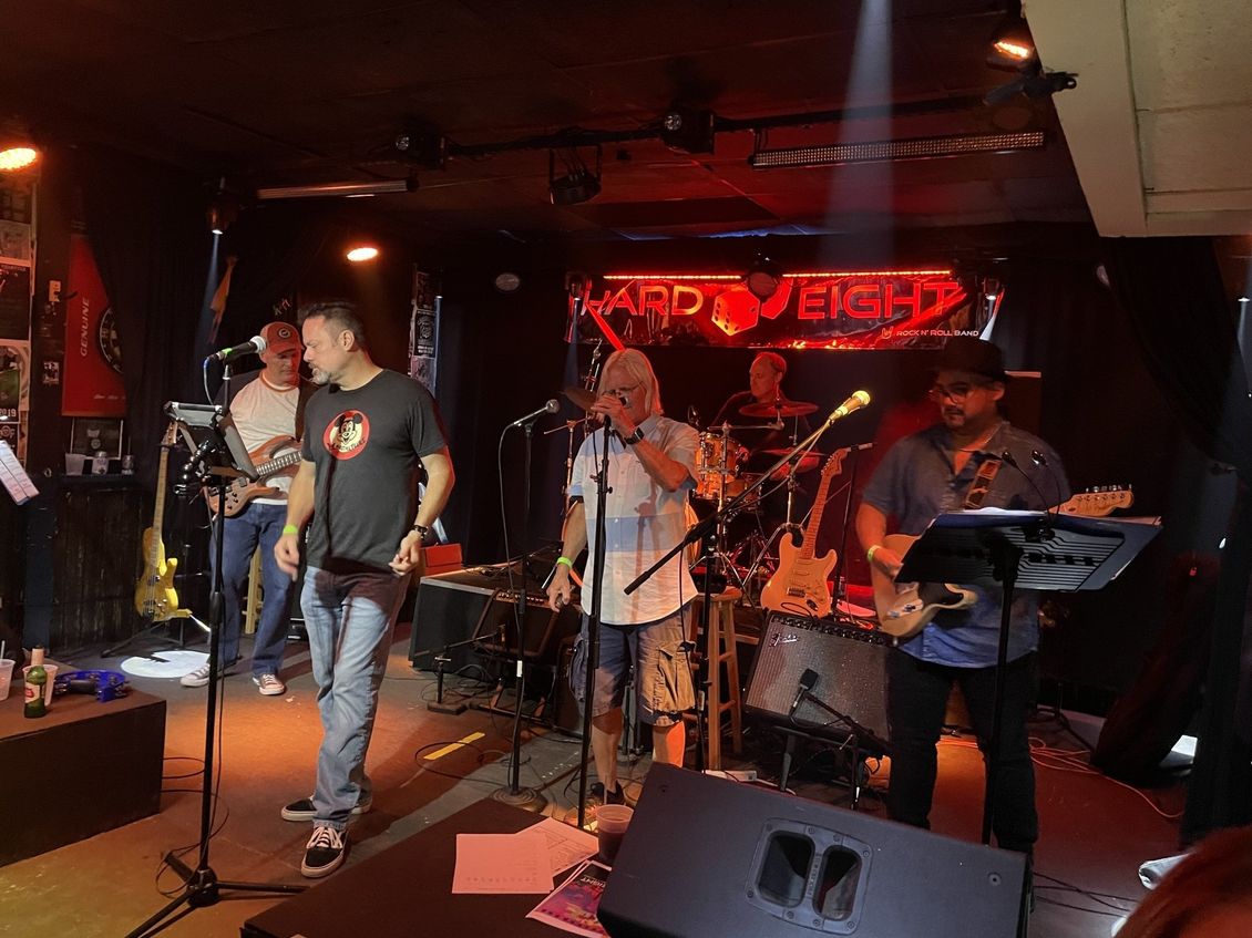 Band performing live on stage at a venue with red lighting; musicians playing instruments, singing.