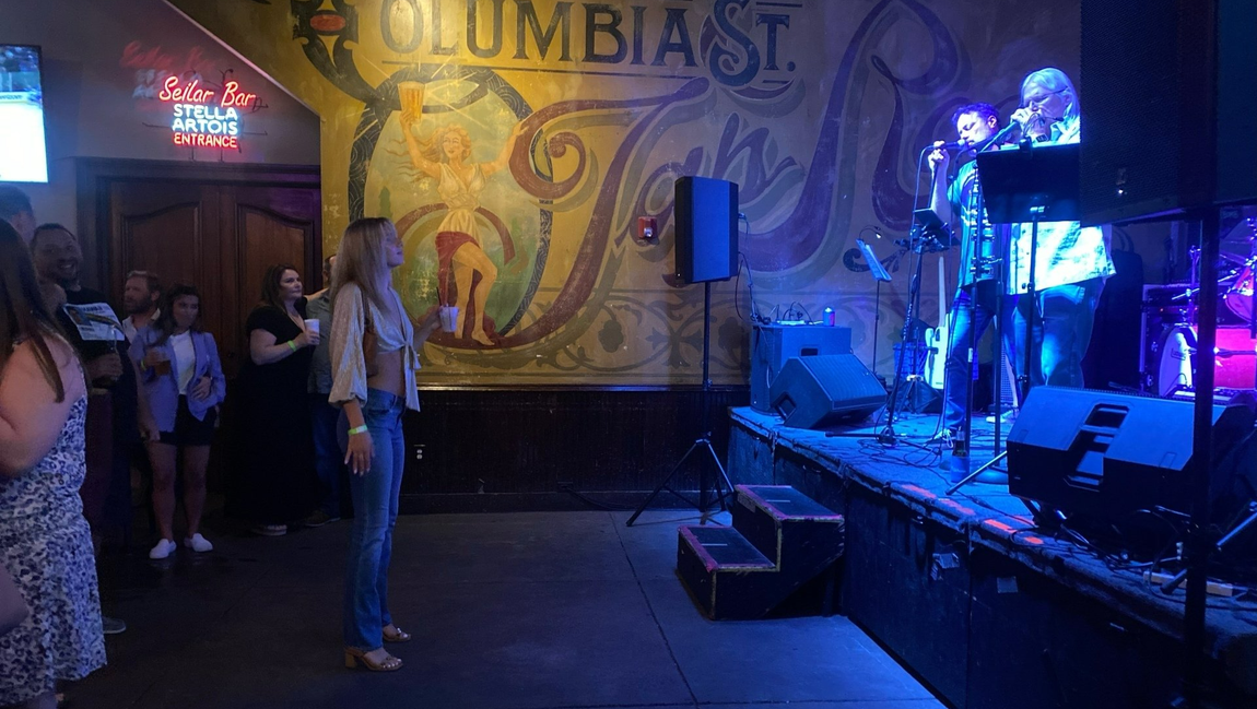 Woman watches band perform on stage at a venue called Columbia St. with mosaic mural backdrop.