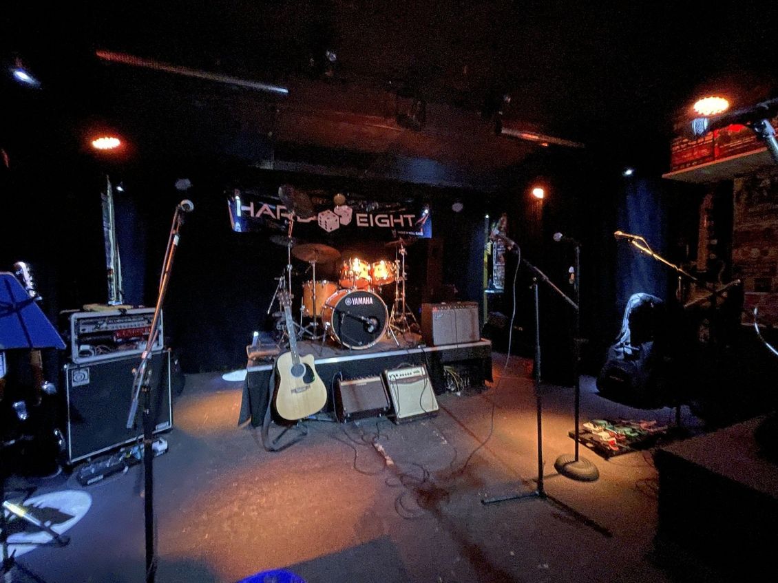 Stage at a music venue. Drums, guitars, amps, and microphones are set up for a performance. Dark interior.
