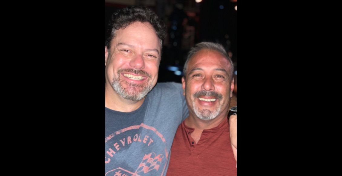 Two smiling men, embracing close together, outdoors at night. One wears a blue shirt, the other a rust-colored shirt.