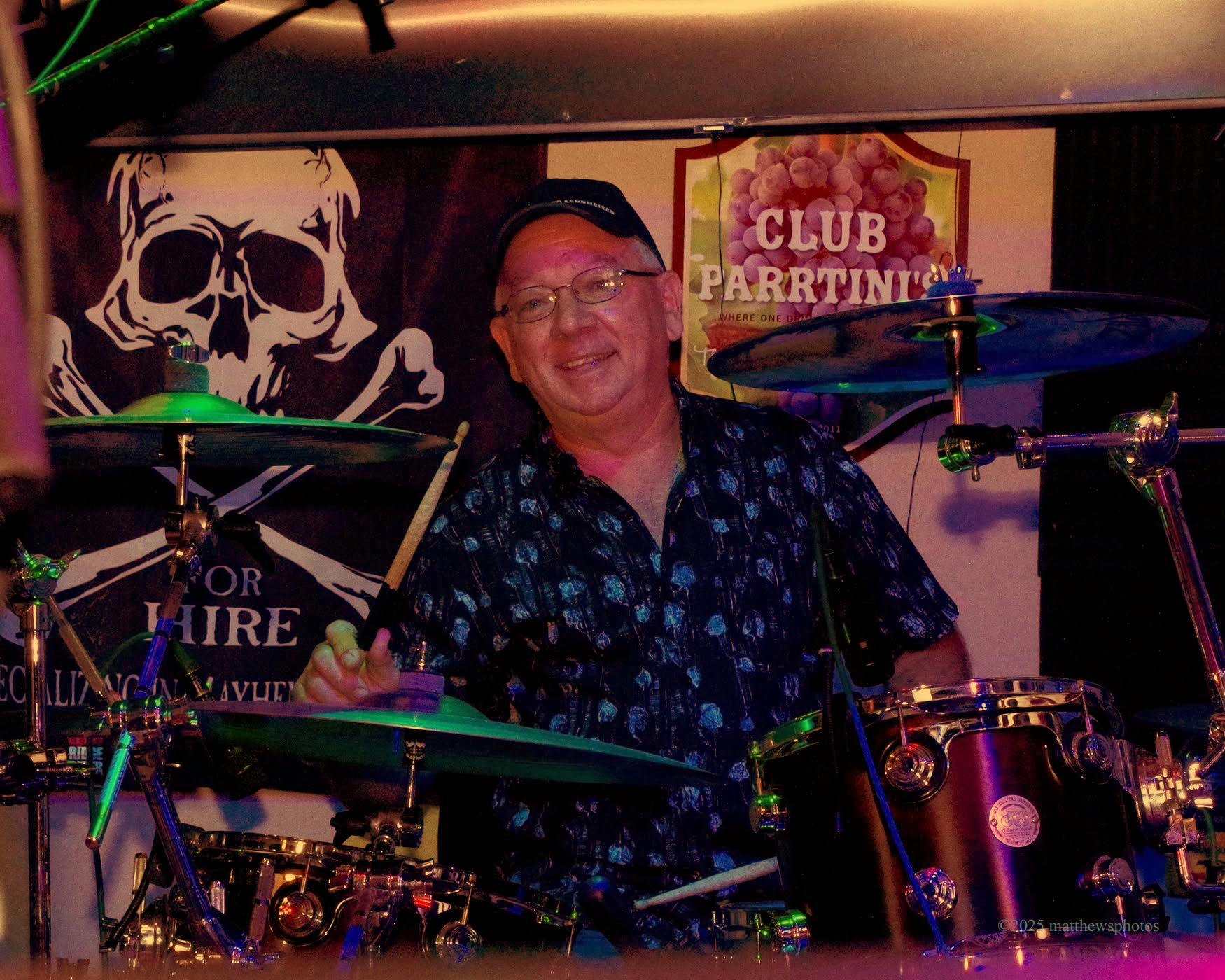Drummer playing a red drum set outdoors, smiling, with drumsticks raised.