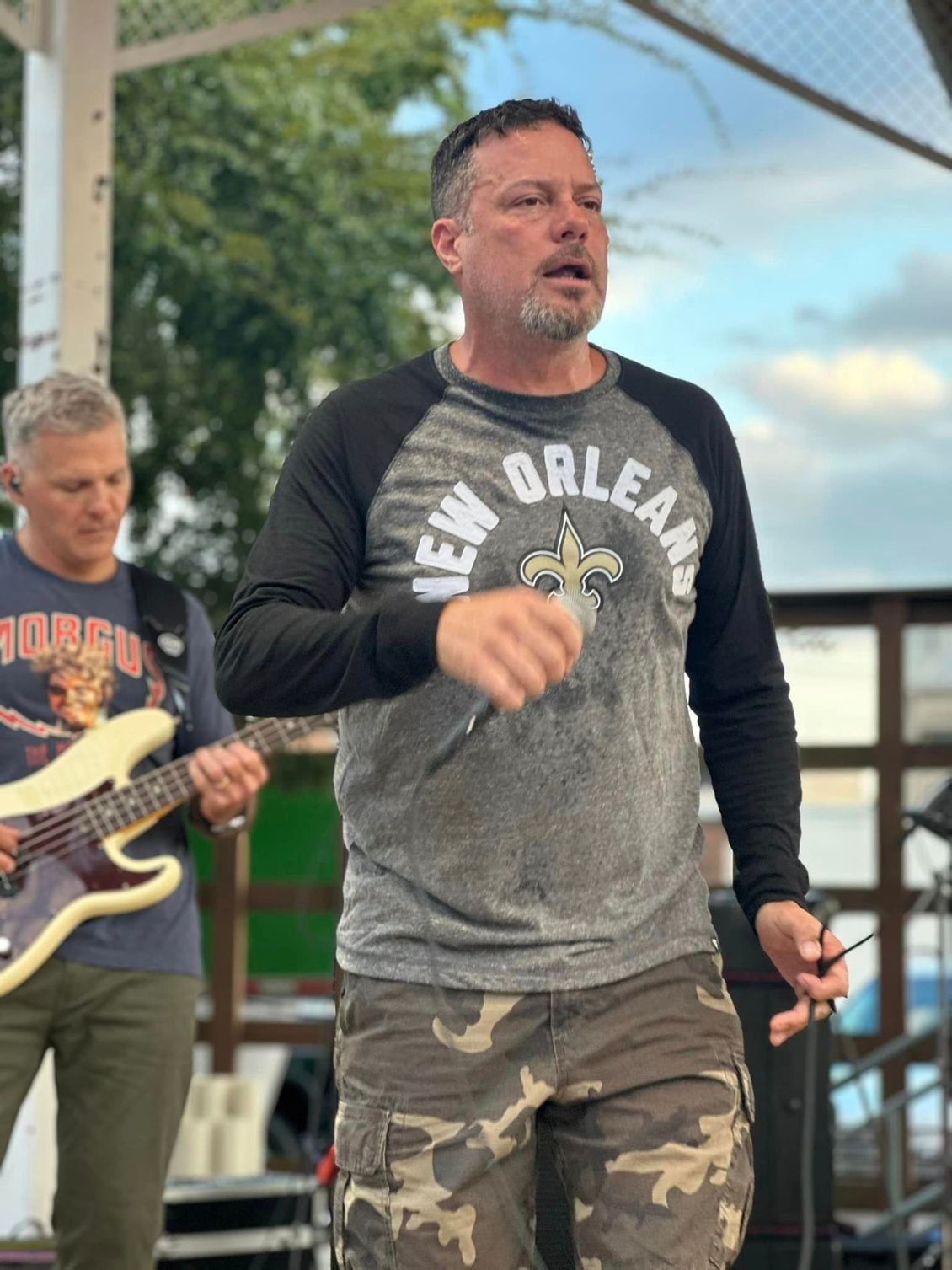 Man singing on stage, wearing a New Orleans shirt and camo pants; guitarist in background.