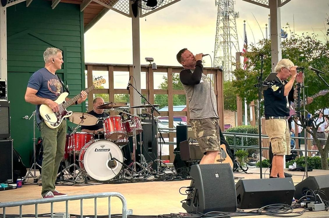 Band performing on an outdoor stage: bass, drums, vocals, and harmonica. Bright light.