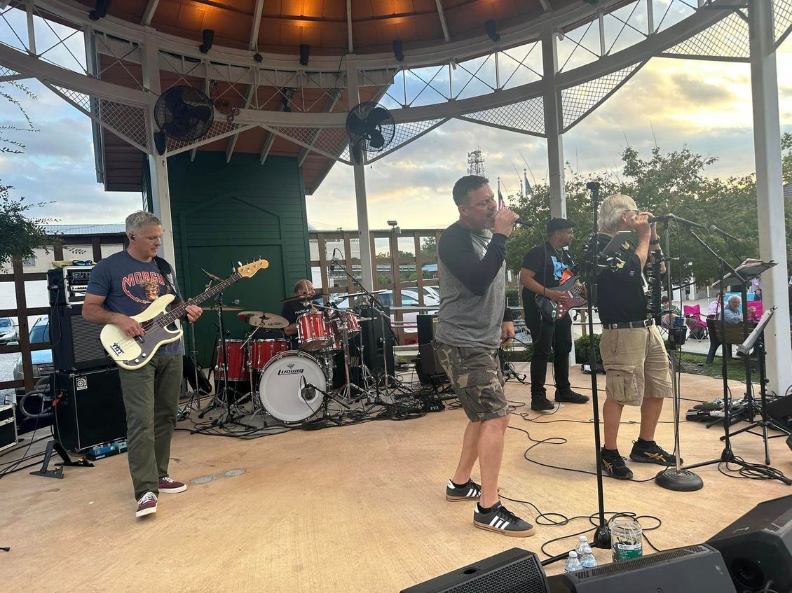 Band performing on a gazebo stage with instruments and microphones; outdoor setting with trees and sky.