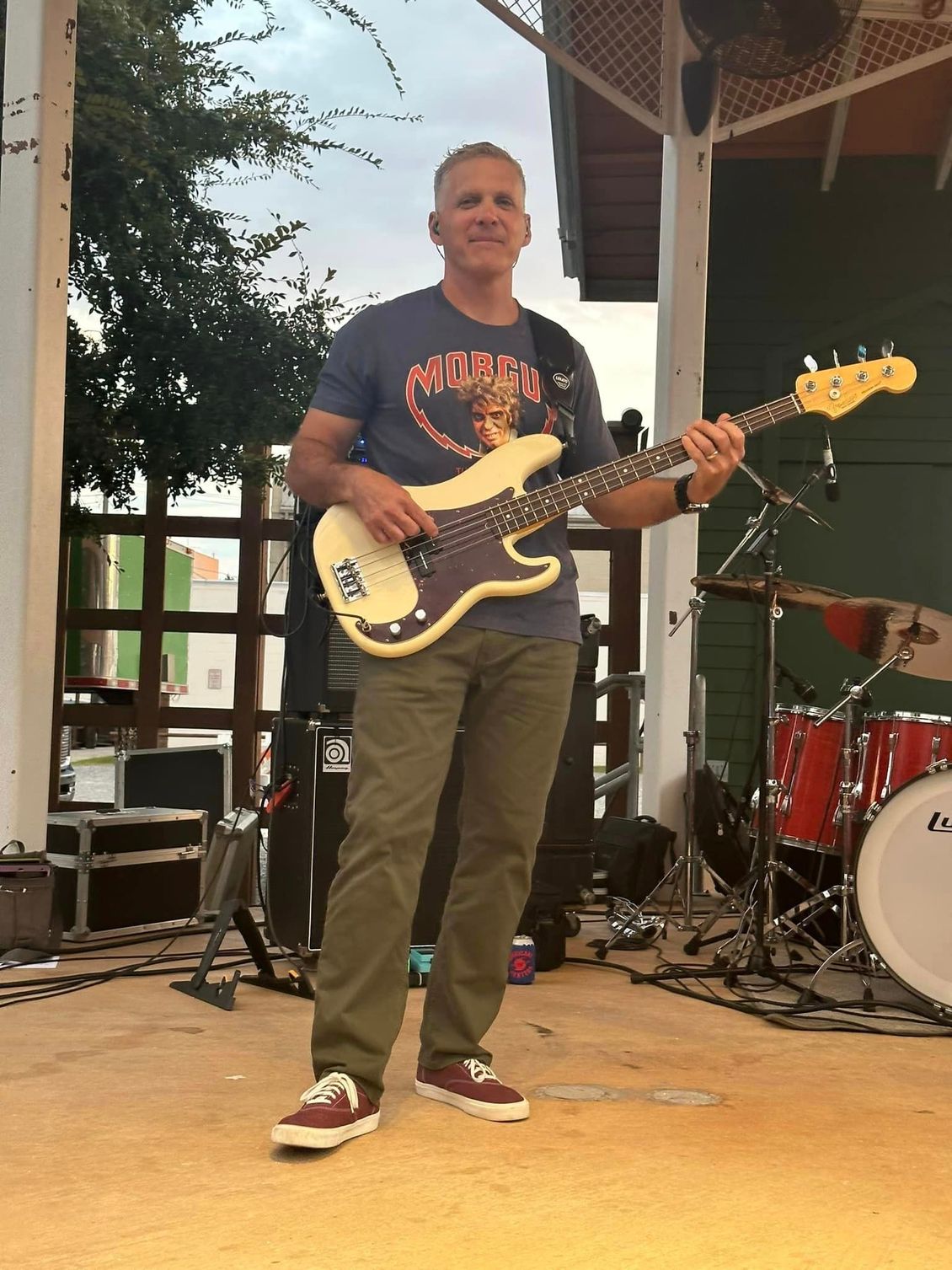 Man playing bass guitar on a stage. He wears a blue shirt, green pants, and red shoes. Drums are behind him.