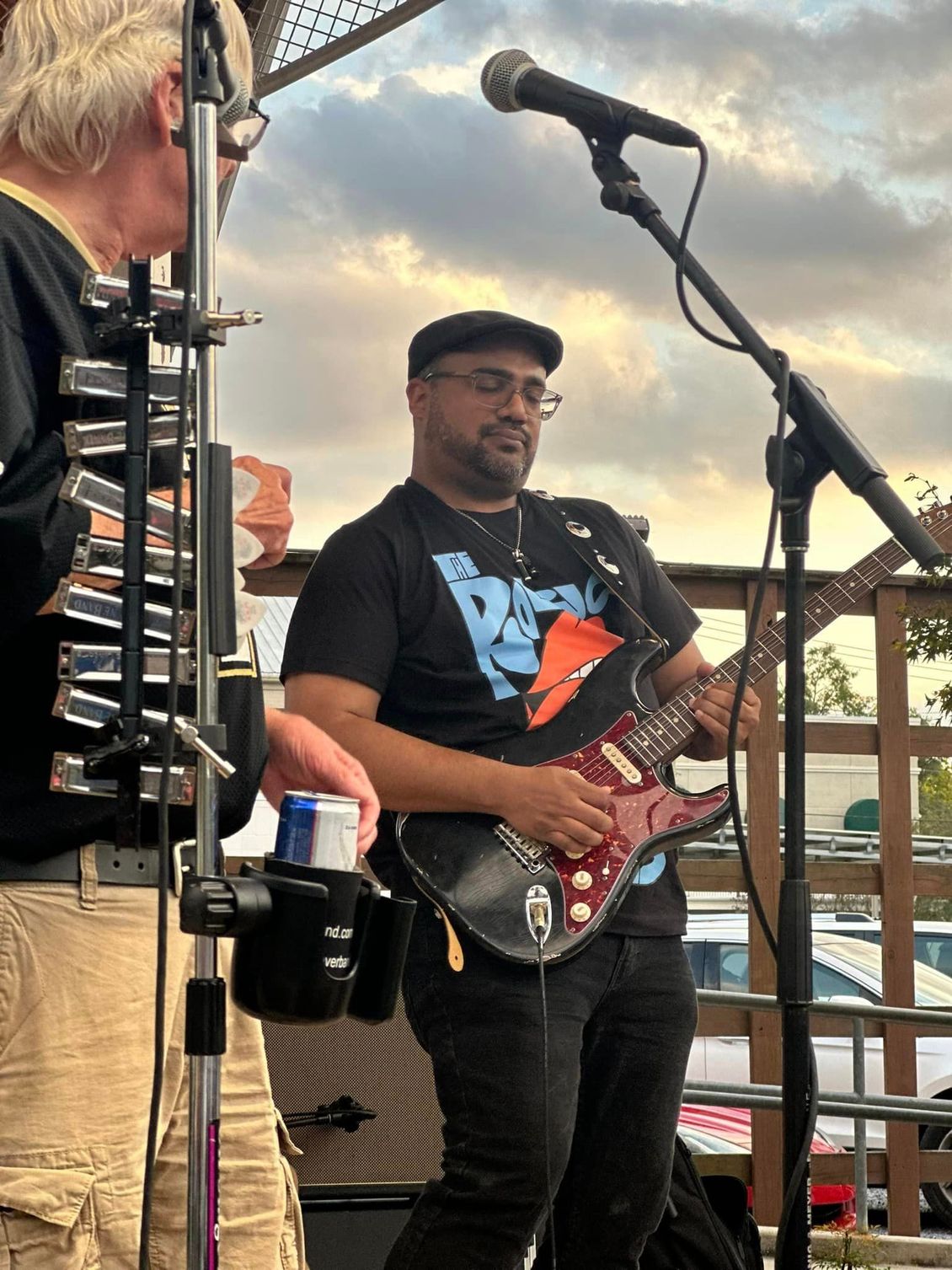 Musician playing guitar, stage, wearing a black shirt and cap. Another musician playing instrument.