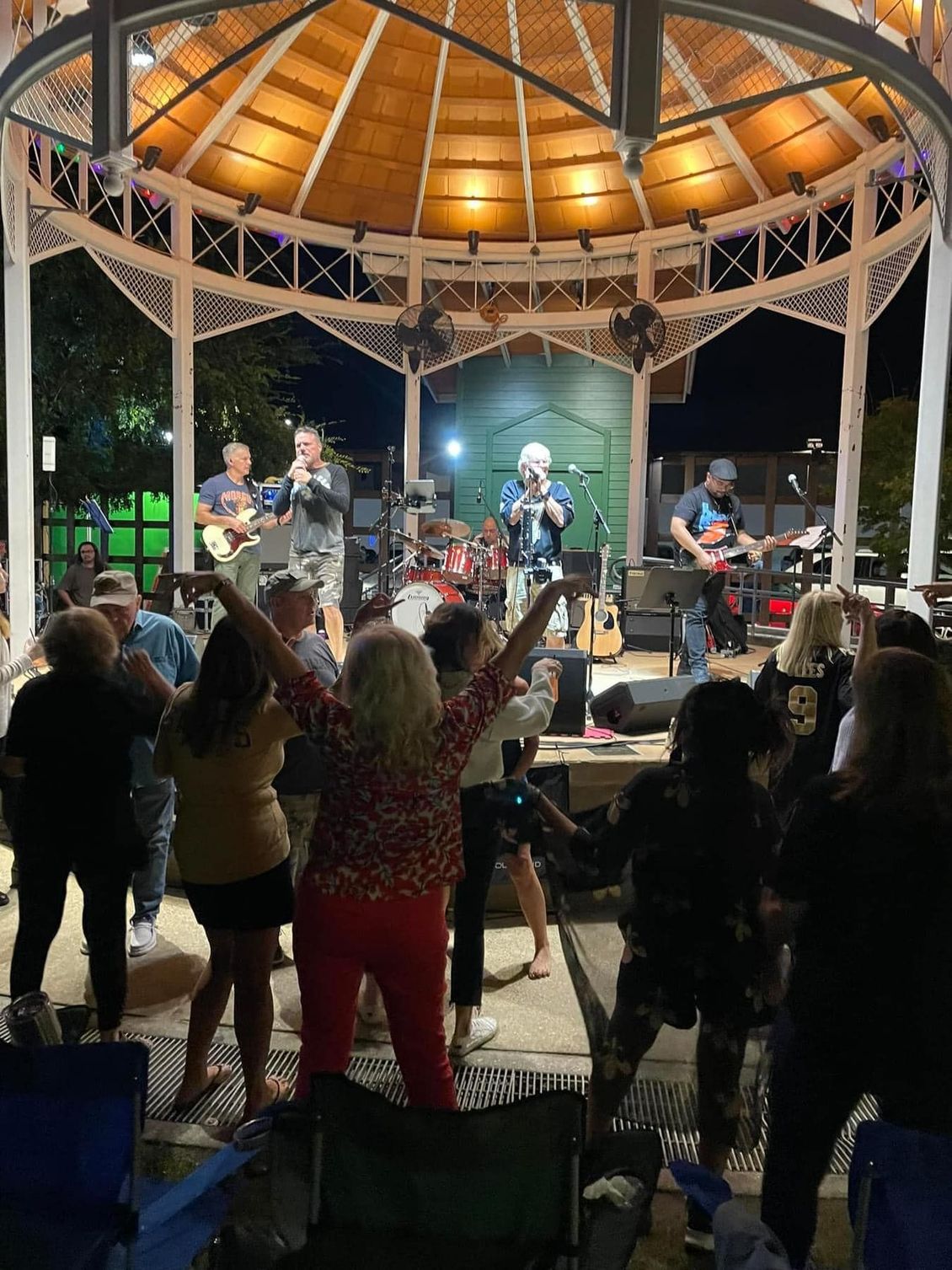 Band performing on stage in a gazebo at night, crowd dancing in front.