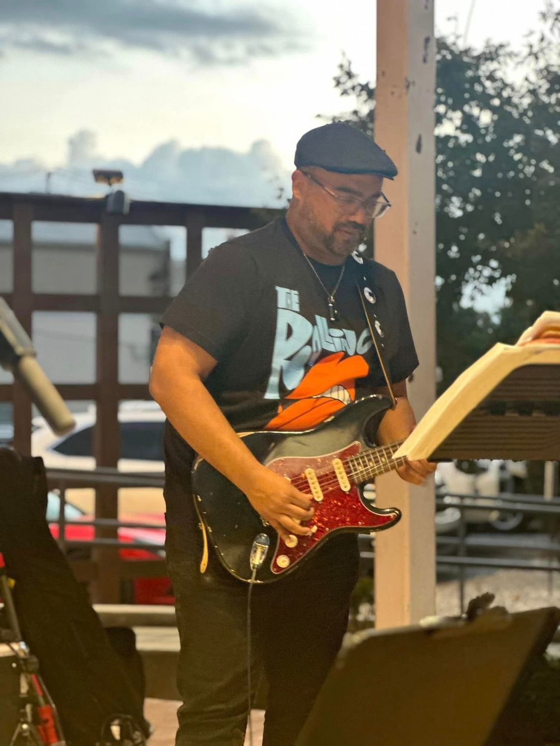 Man playing a black electric guitar with a red pickguard, outdoors. He wears a cap and glasses.