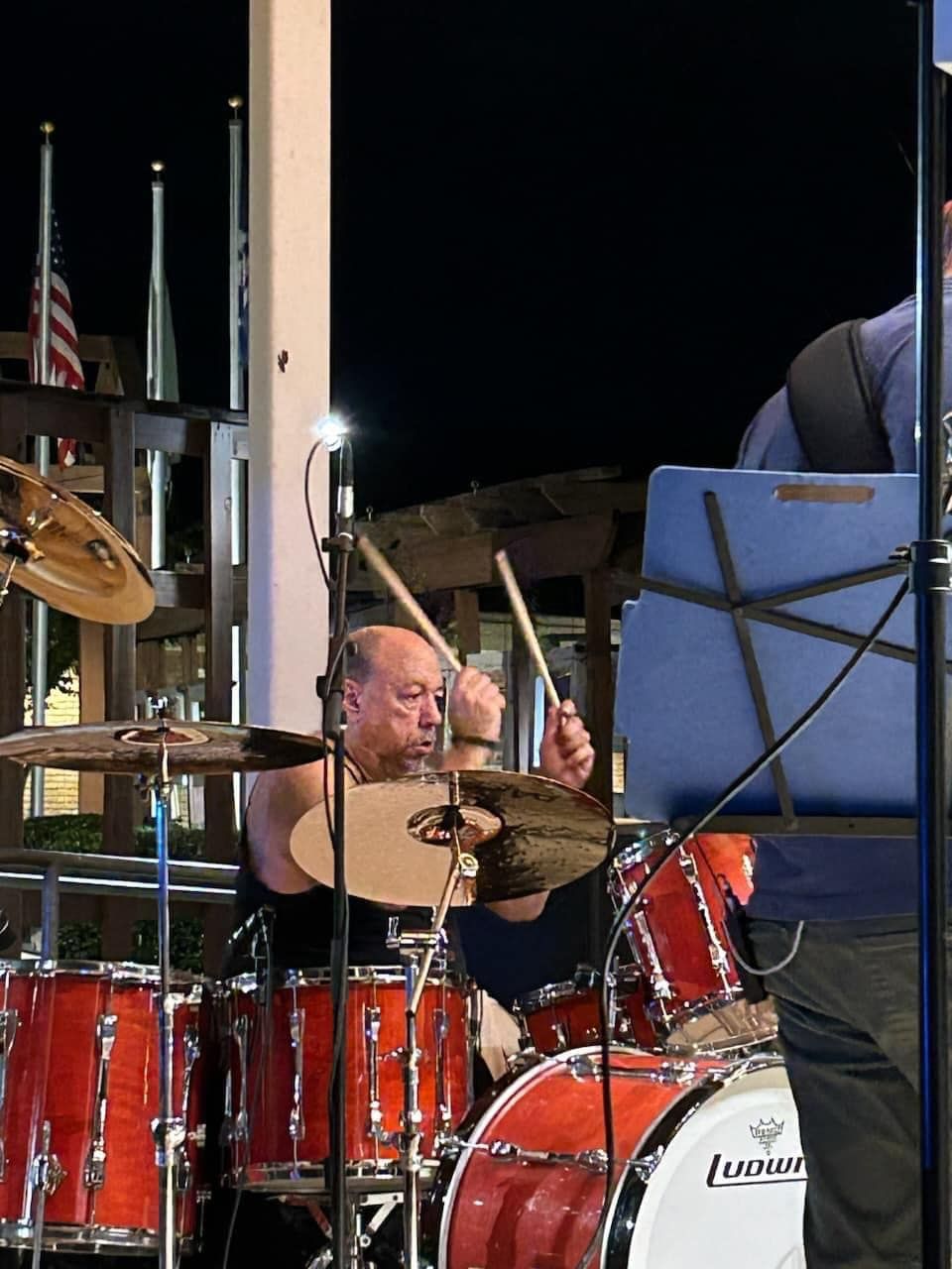 Drummer playing red drum kit outdoors at night, under a white pillar.