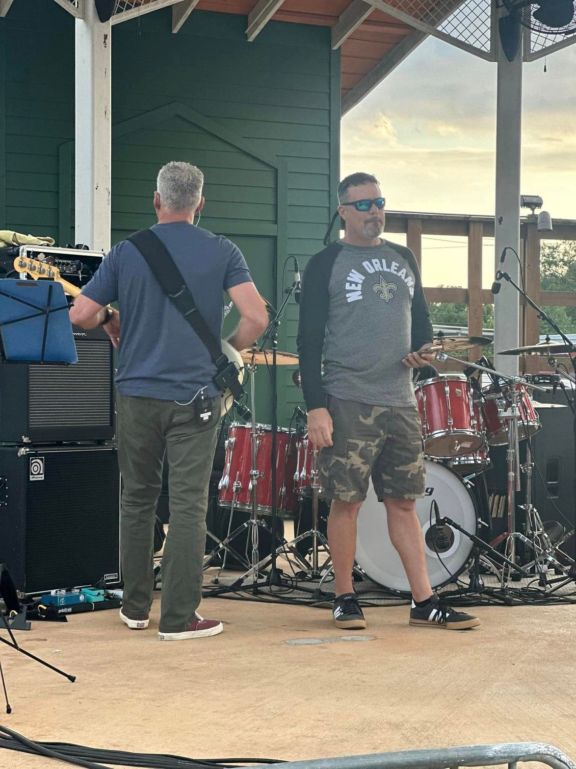 Two men on a stage, one playing guitar, the other in sunglasses. Drums in the background. Outdoor setting.
