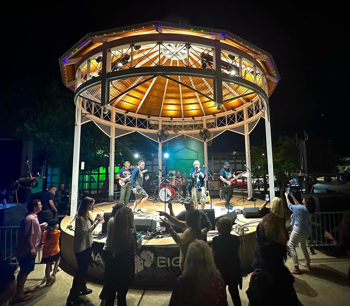 Band performing on a lit-up gazebo stage at night. Crowd cheers in front.