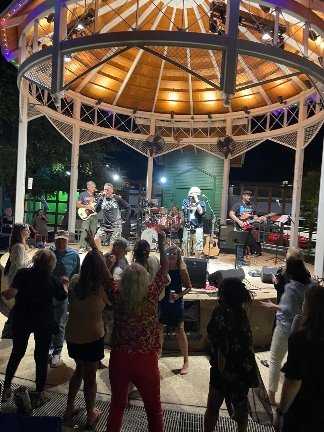 Band playing on gazebo stage; people dancing in front. Nighttime setting, overhead lights.