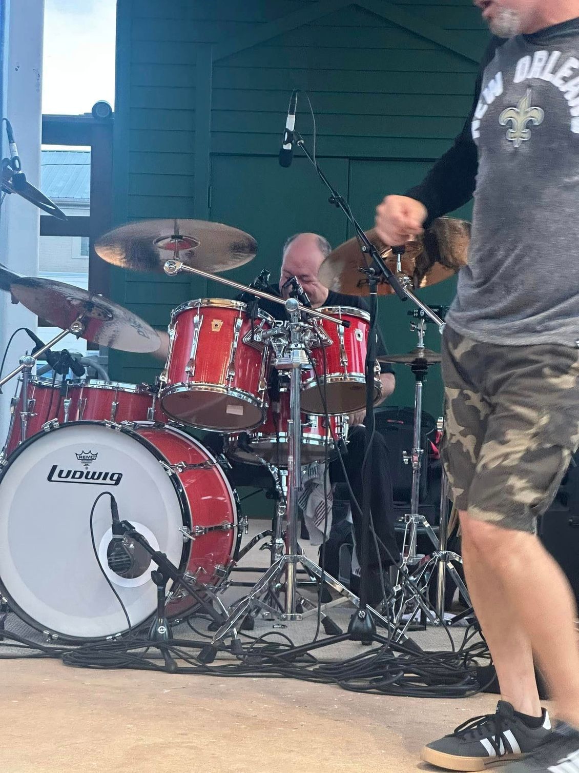 Drummer playing a red drum set onstage; a man in camouflage shorts moves near him.