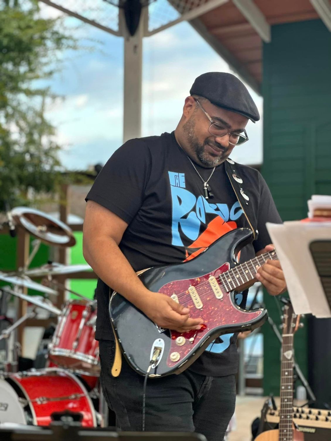 Man playing electric guitar outdoors. Wearing black cap, shirt with blue graphic. Drum set visible.