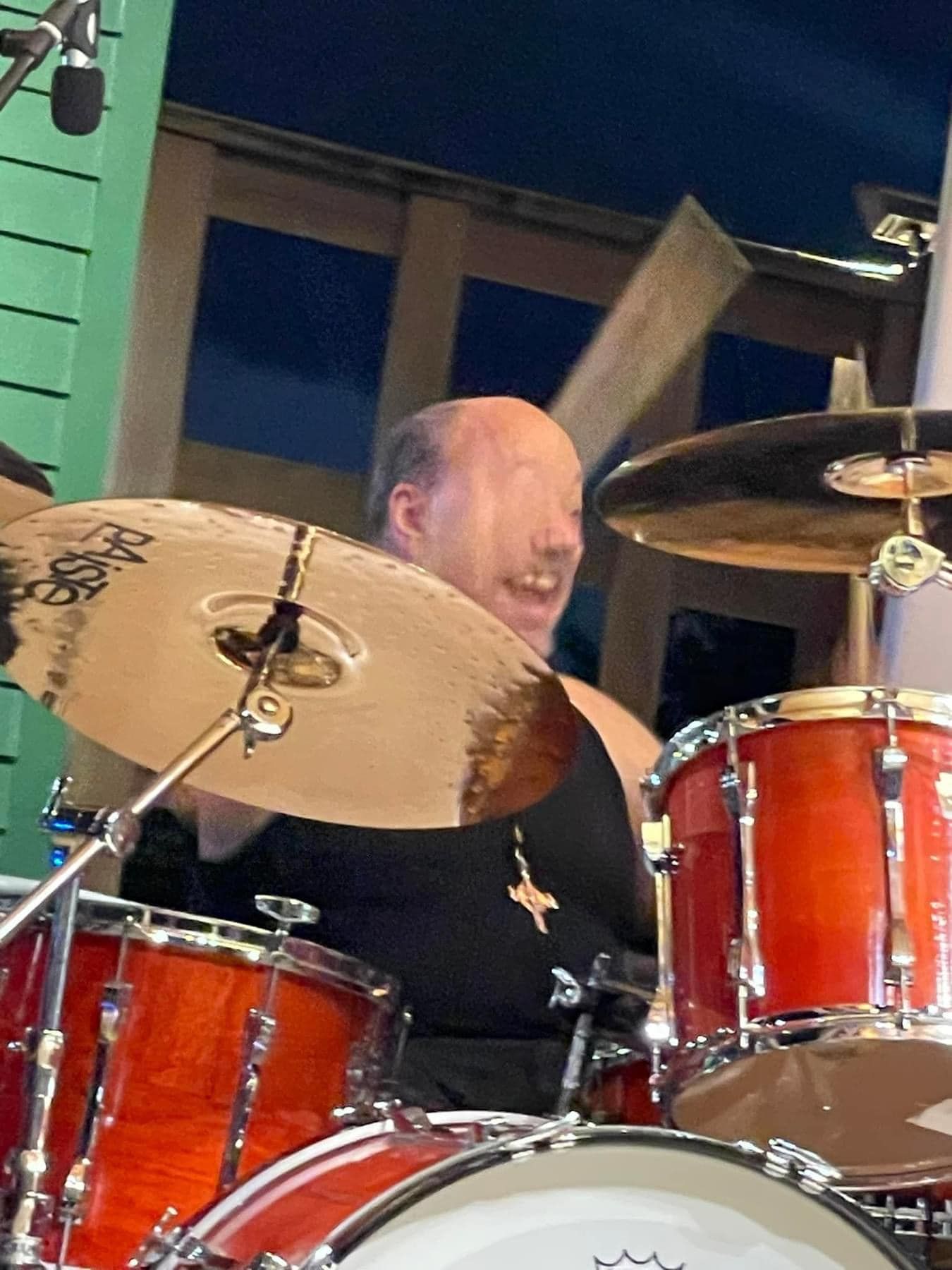 Drummer playing a drum set outdoors; reddish drums, brass cymbals, smiling expression.