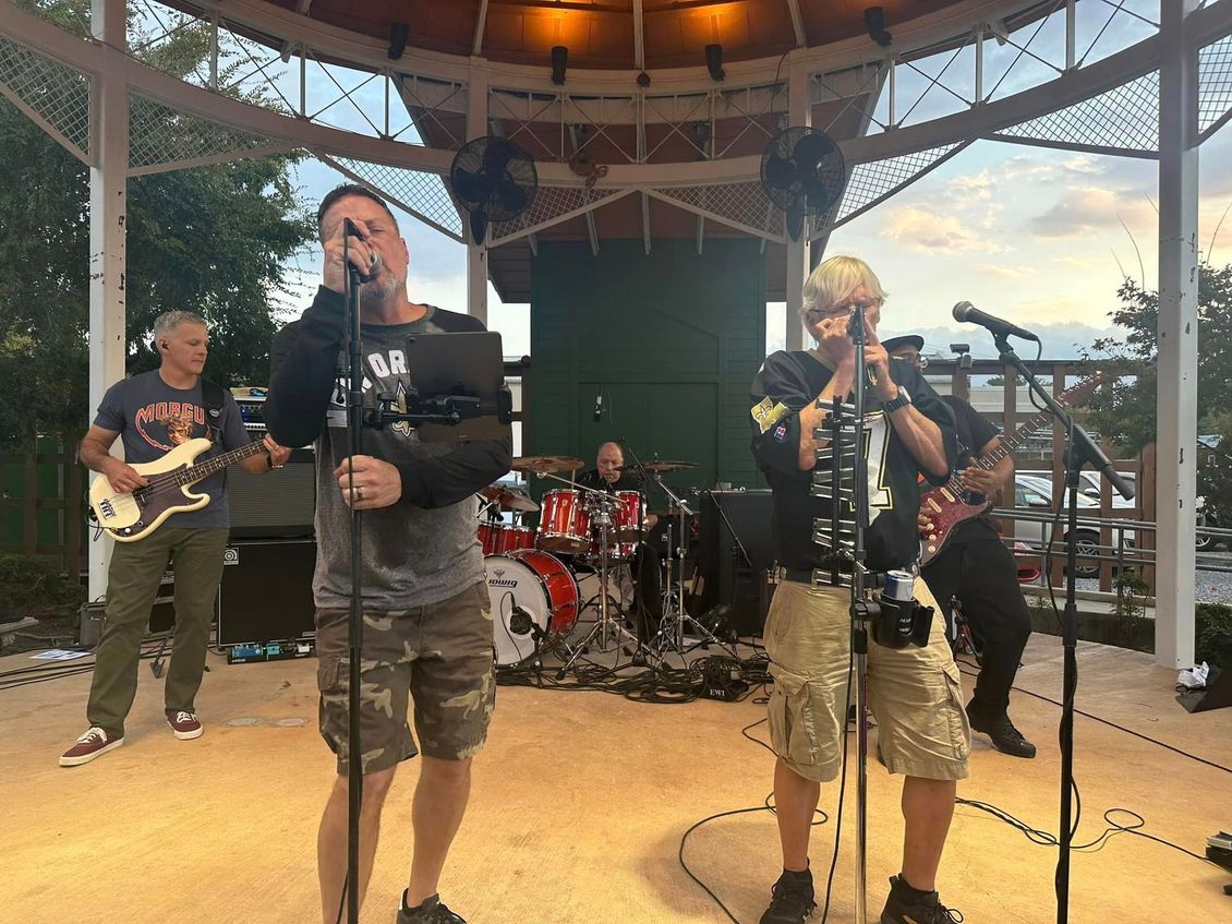 Band performing on a gazebo stage; two vocalists, bassist, drummer. Outdoor setting, overcast sky.