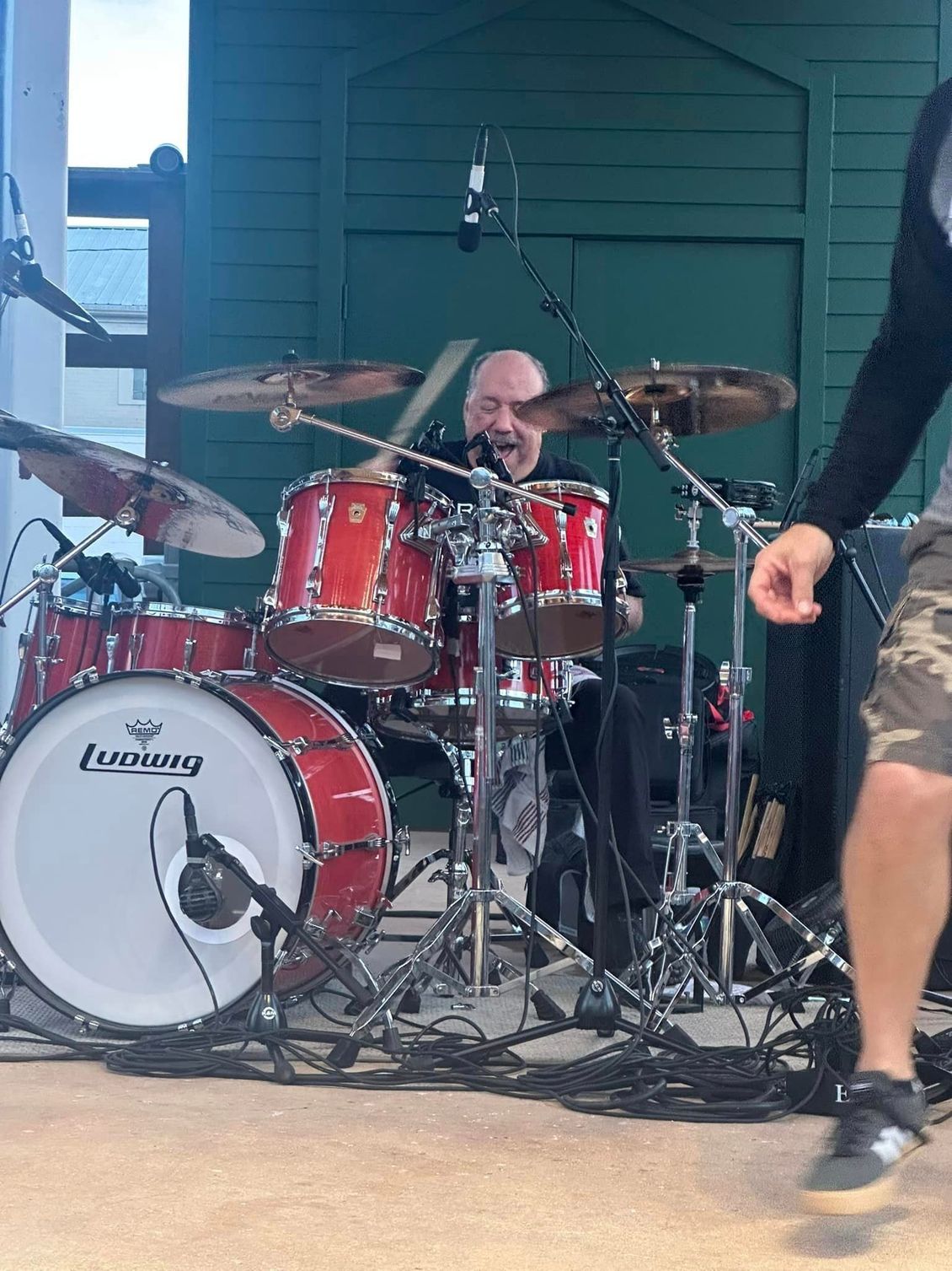 Drummer playing a red drum kit on a stage, smiling.