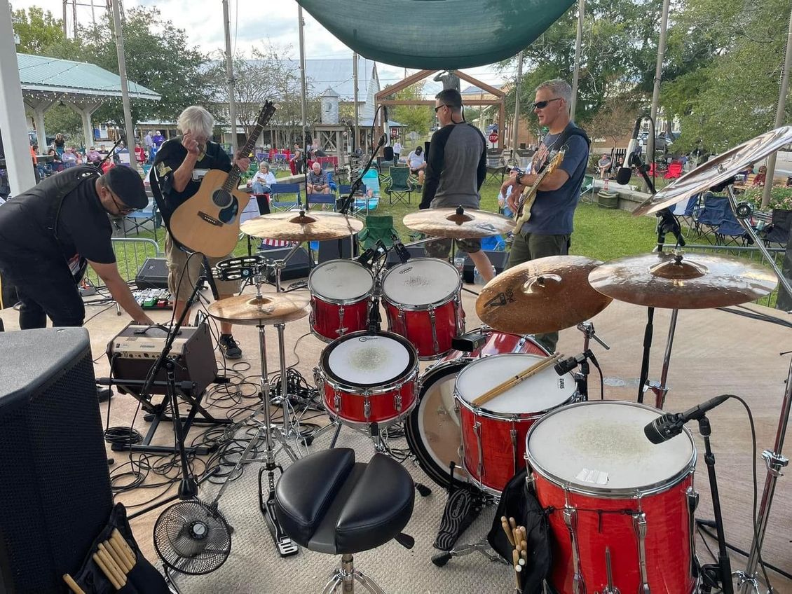 A band performs outdoors: drummer, guitarist, vocalist, on stage with crowd in background.