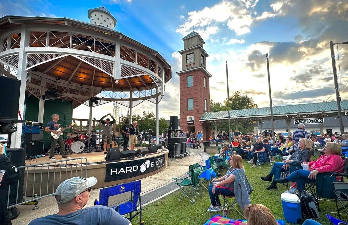 Band performing on stage in a gazebo, crowd watching, clock tower in the background, outdoor event.