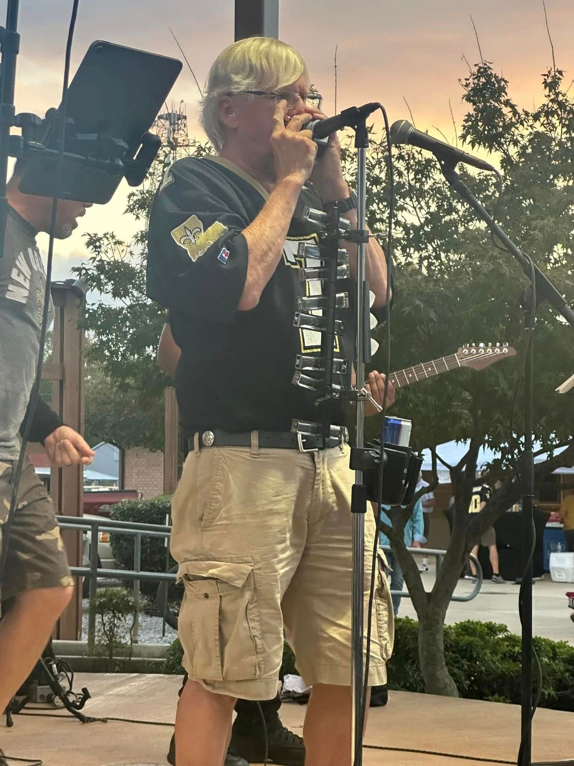 Man playing harmonica and guitar on outdoor stage, wearing a black shirt and khaki shorts.