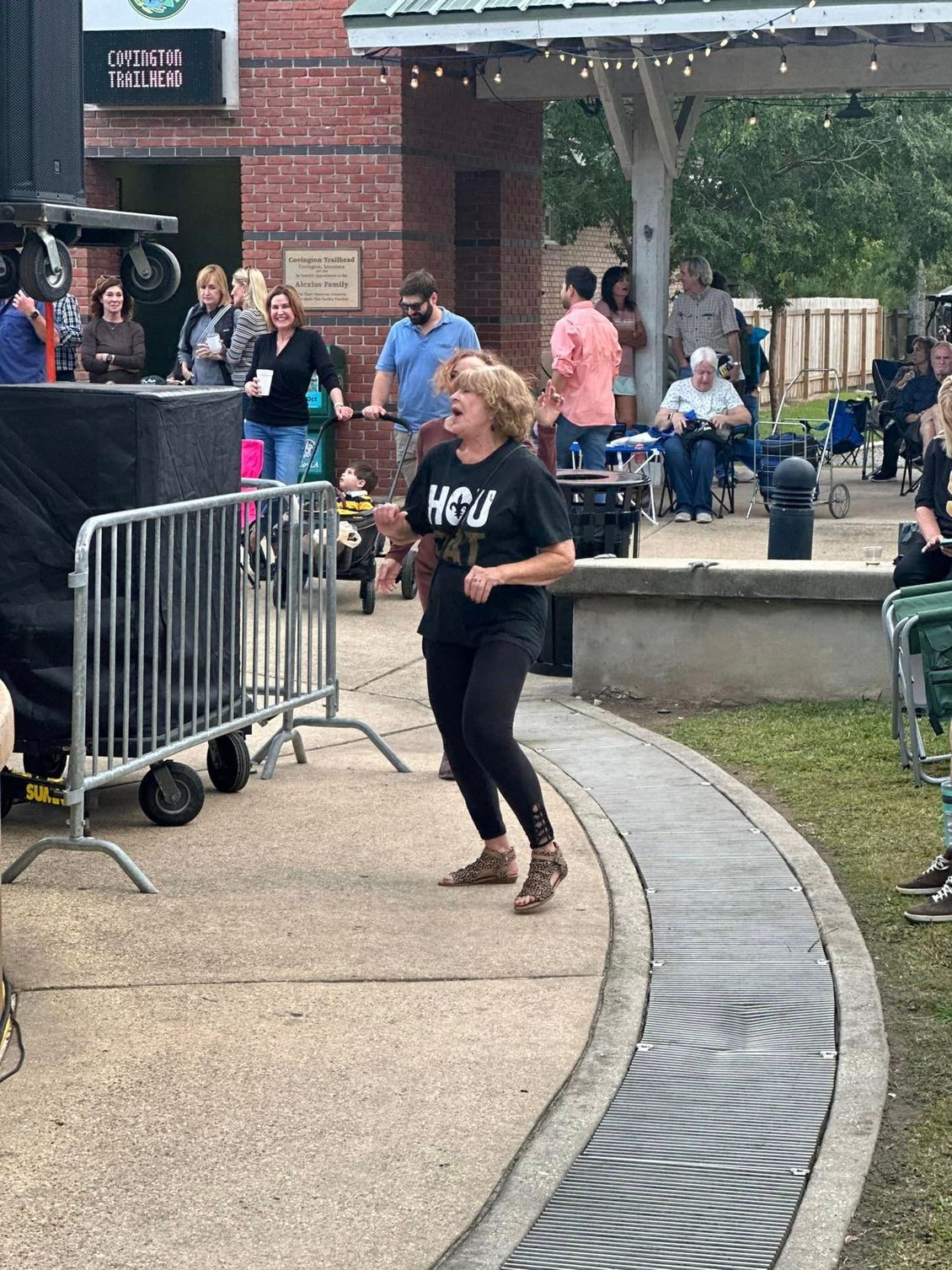 Woman dancing near a stage at an outdoor event, people in background, brick building.