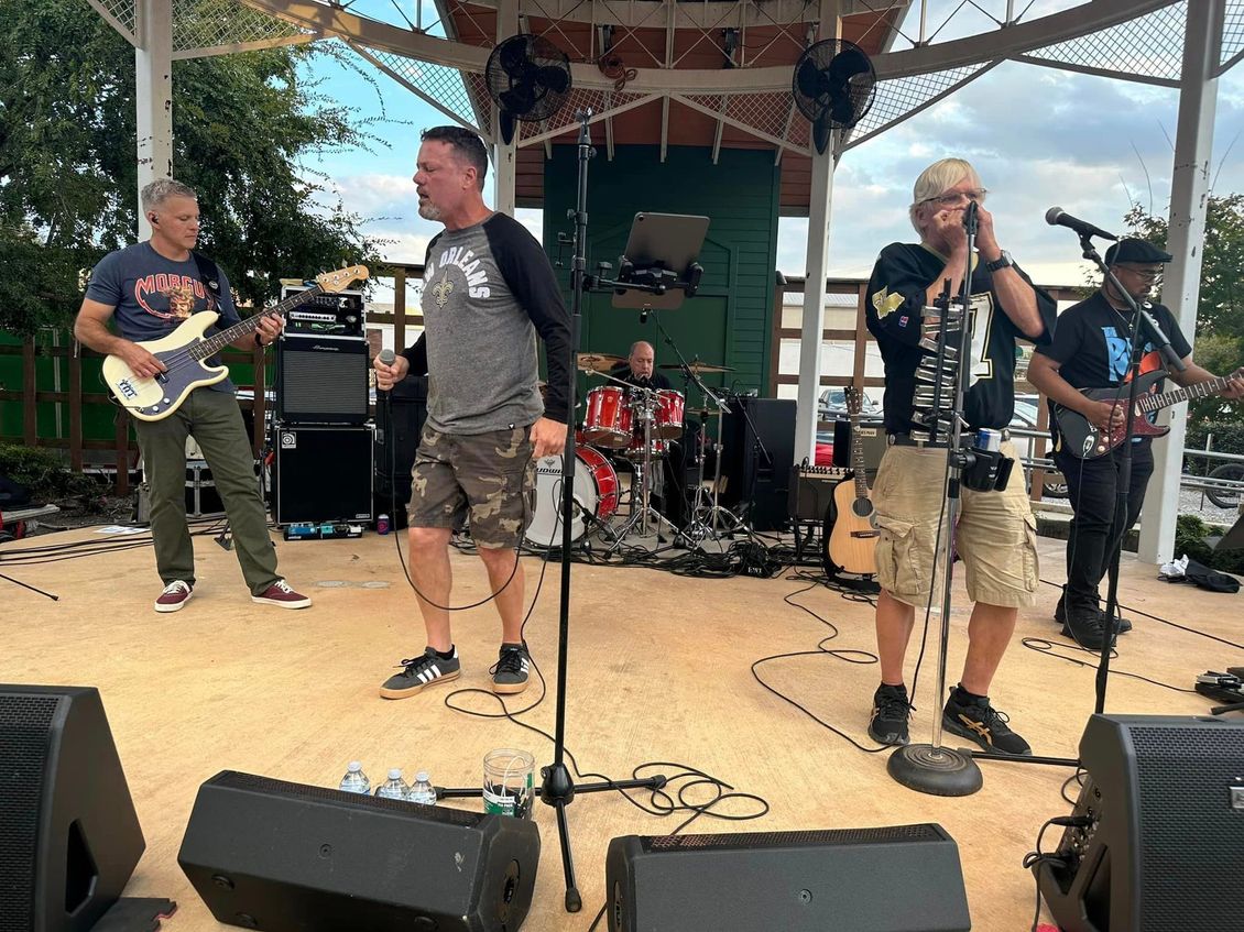Band playing on an outdoor stage: guitar, bass, drums, and vocalist with harmonica.