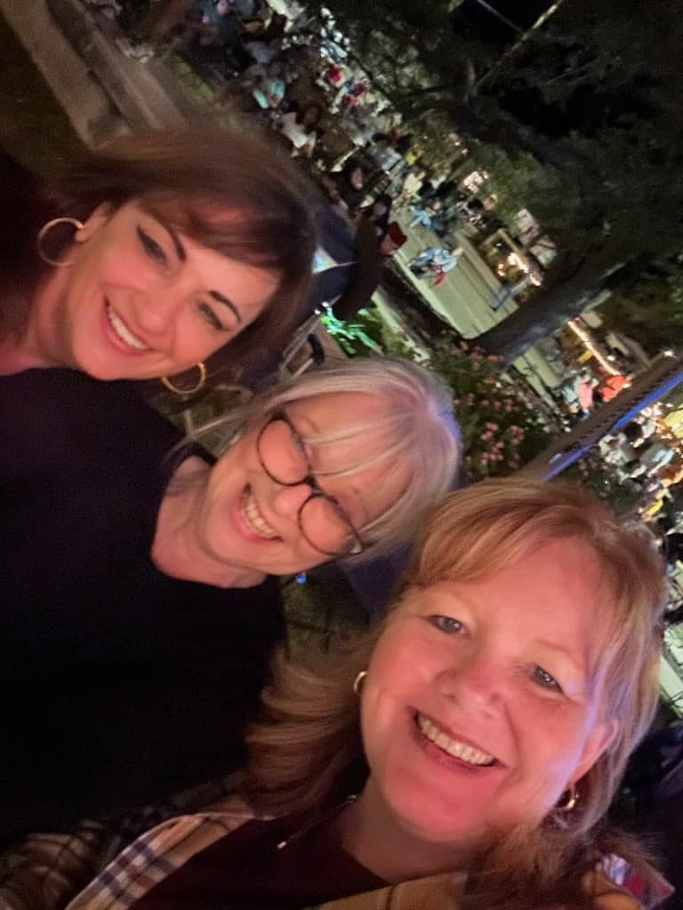 Three smiling women pose for a selfie, illuminated at night. Background is a blurry event setting with lights.
