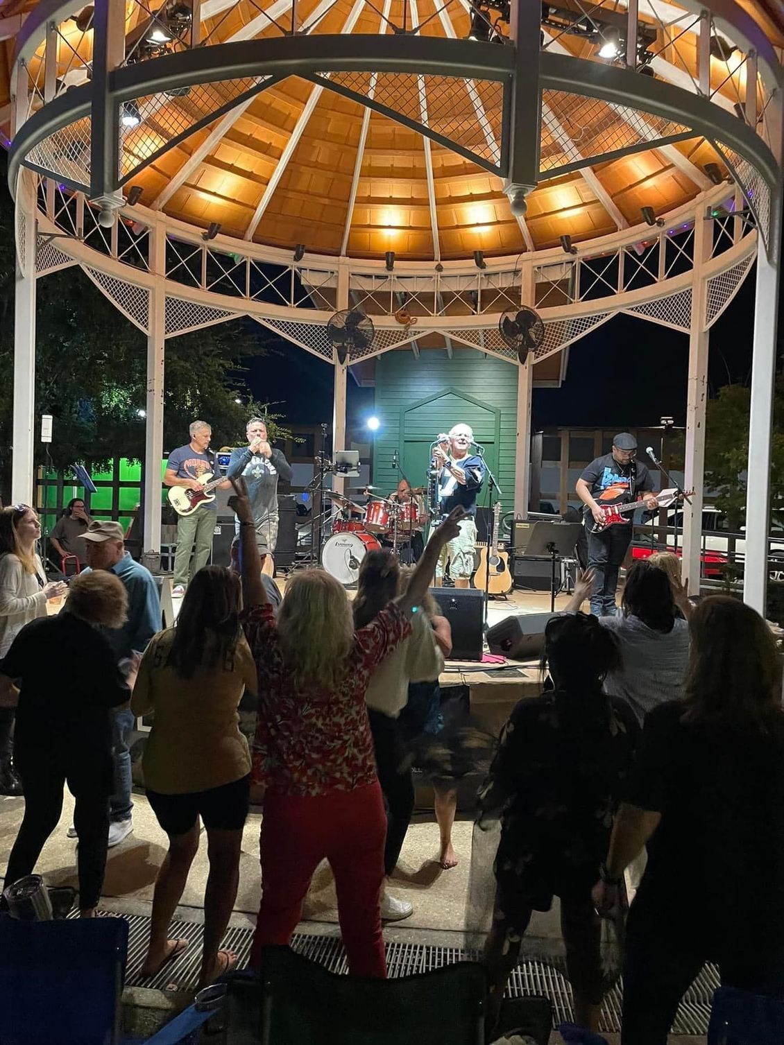 Band performs under gazebo; crowd dances. Night scene with lights.