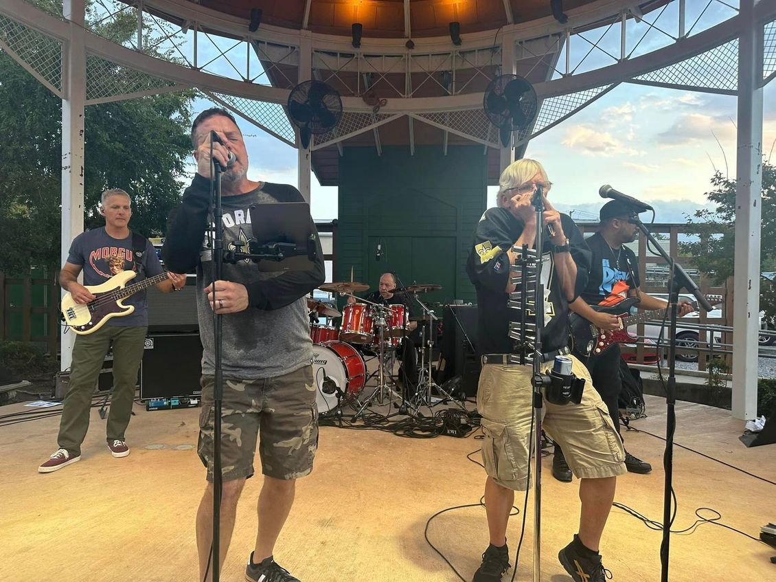 Band performing on outdoor stage, singing into microphones. Drummer in the back, guitarist, bassist, and two other singers.