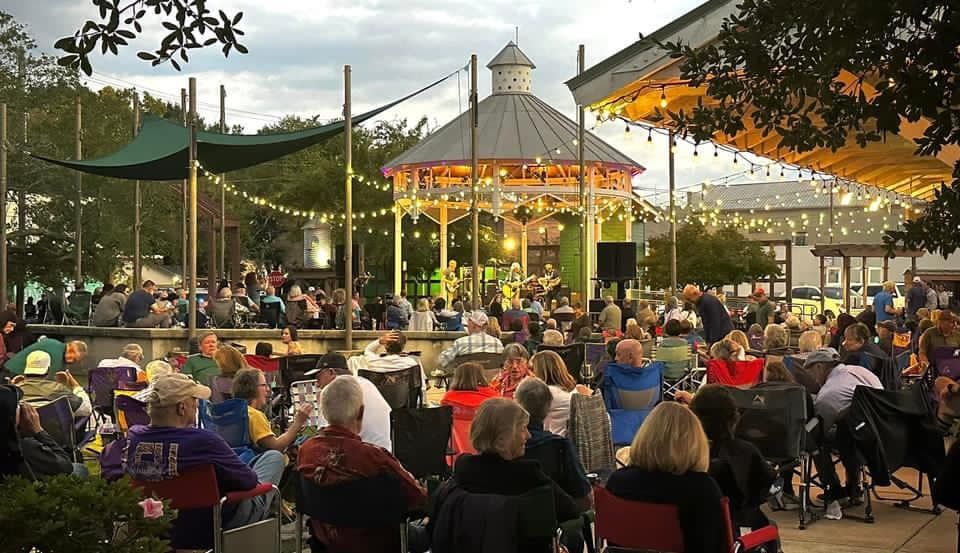 Crowd at outdoor concert; gazebo stage with band, strung lights, seated audience with chairs.