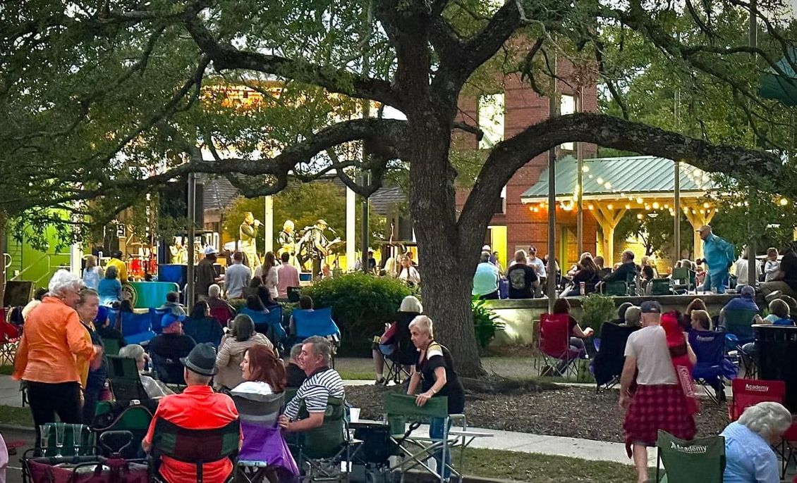 People gathered outdoors, sitting in chairs, watching a performance on a stage in a park at dusk.