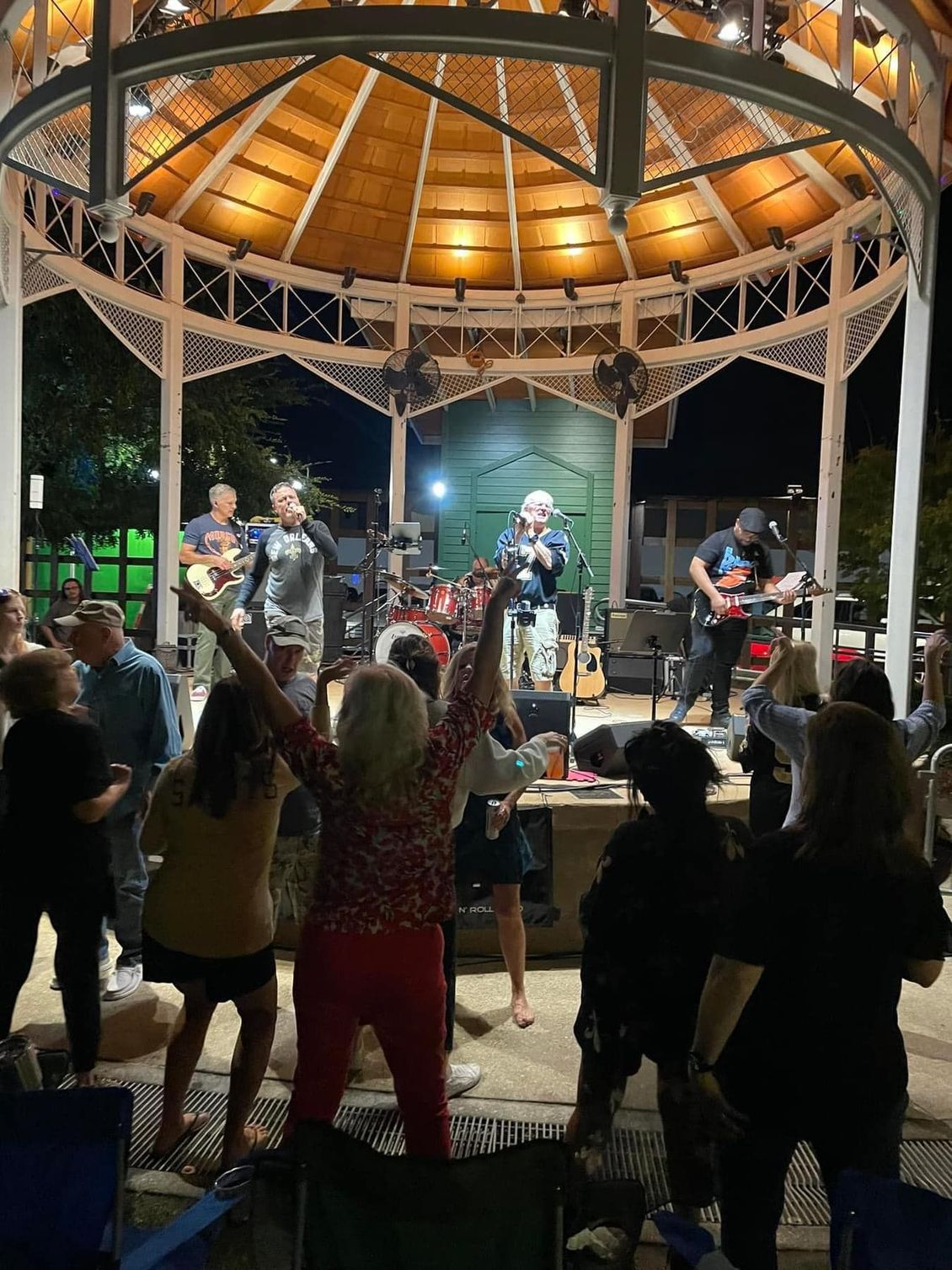Band performing on stage in a gazebo at night, people dancing and enjoying the show.