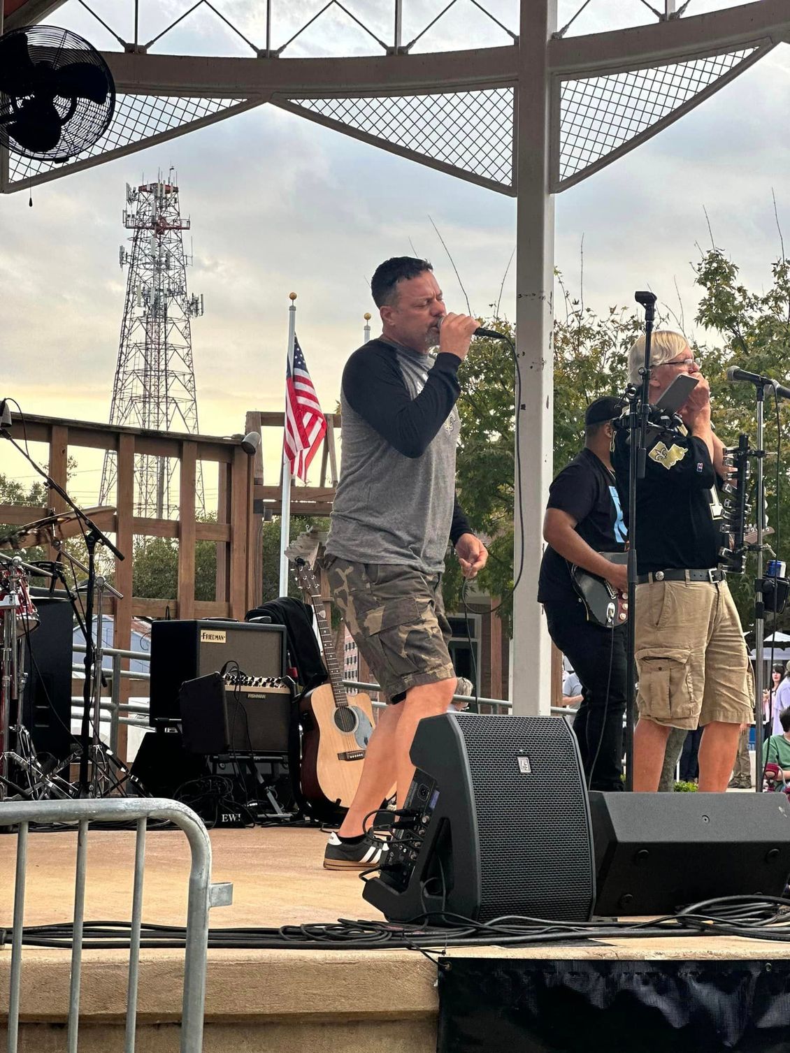 A band performs on an outdoor stage. Lead singer in gray shirt sings; others play instruments.