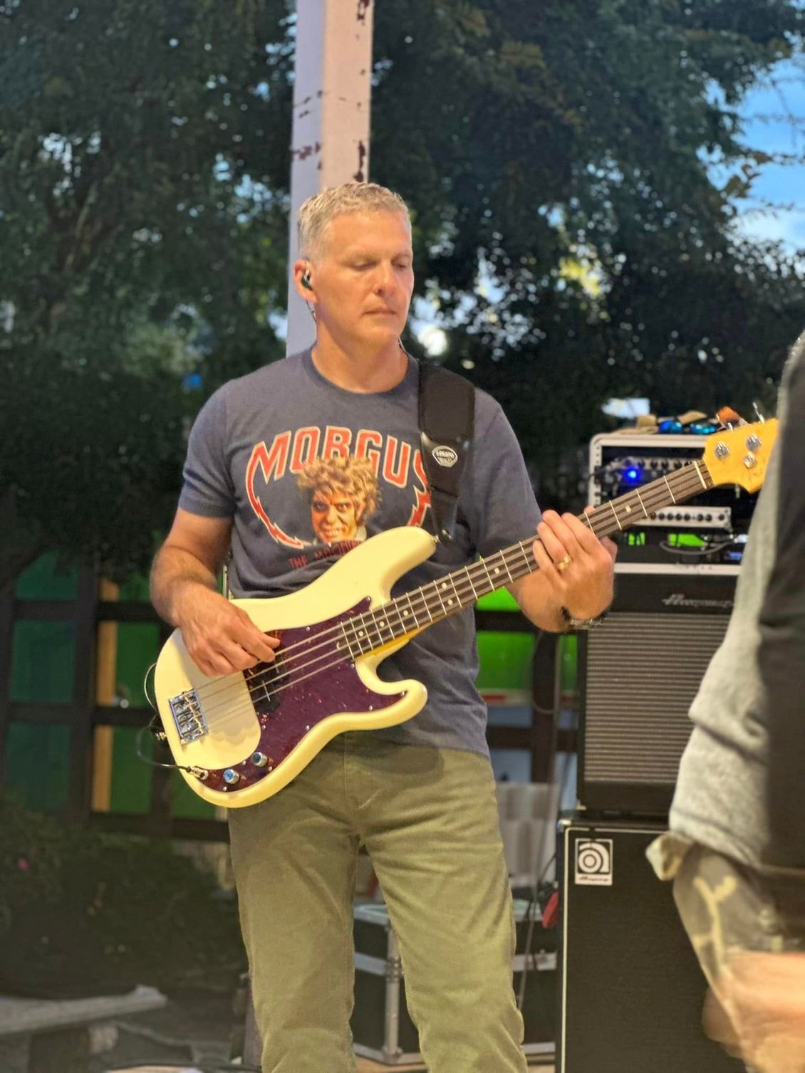 Man playing bass guitar outdoors, wearing a blue t-shirt and green pants.