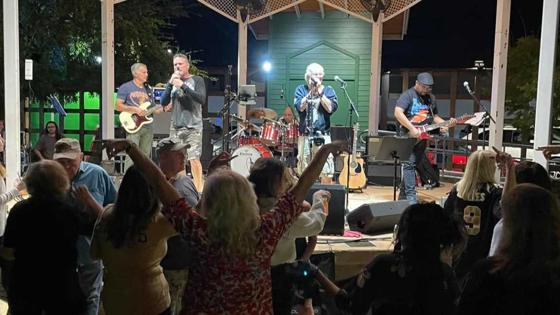 Band performing on outdoor stage, people dancing in front. Nighttime setting with lights.