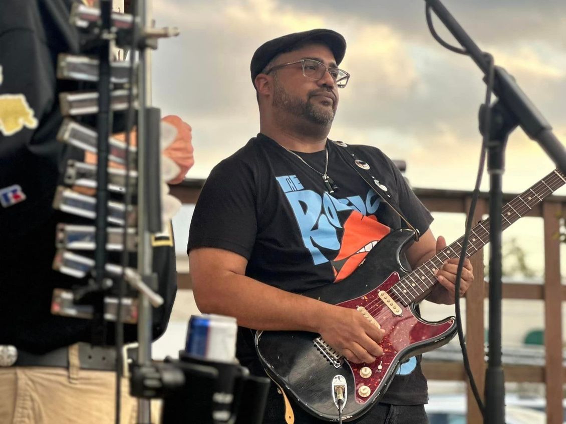 Man plays a red and black electric guitar on stage, wearing a black cap and t-shirt with a blue graphic.