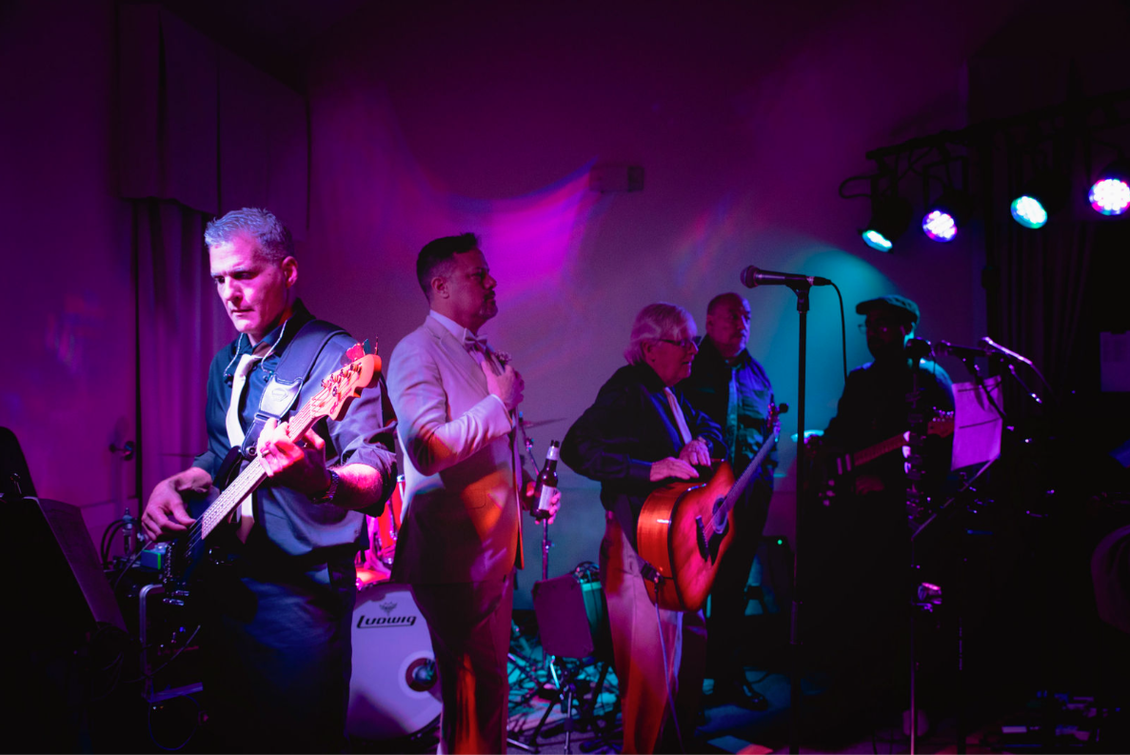 Band performing on a stage with purple and blue lighting. Musicians playing bass, singing, and playing guitar.
