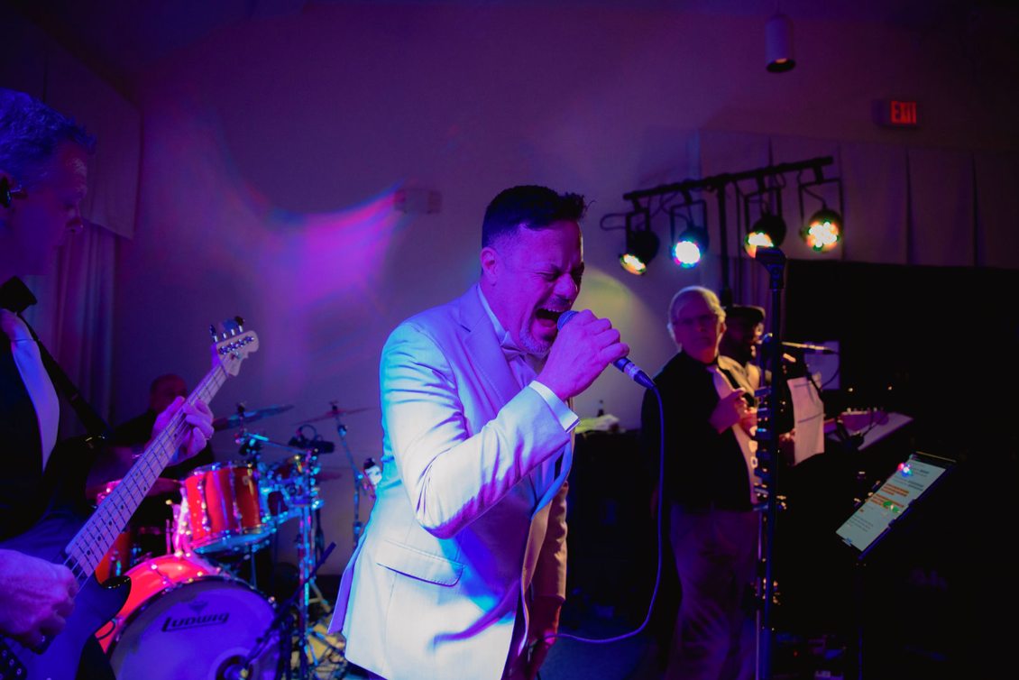 Band performing live on stage under blue and purple lights. Singer in white jacket singing into a microphone.