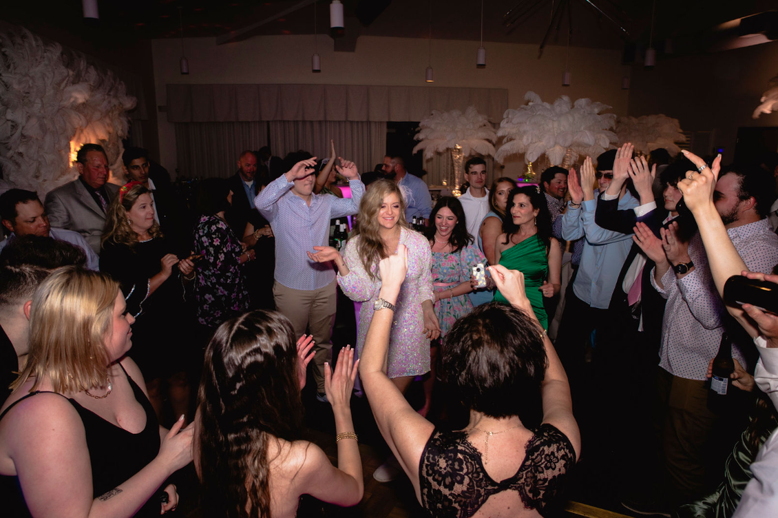 People dancing and celebrating in a well-lit room. Some have hands raised. White decor on walls.