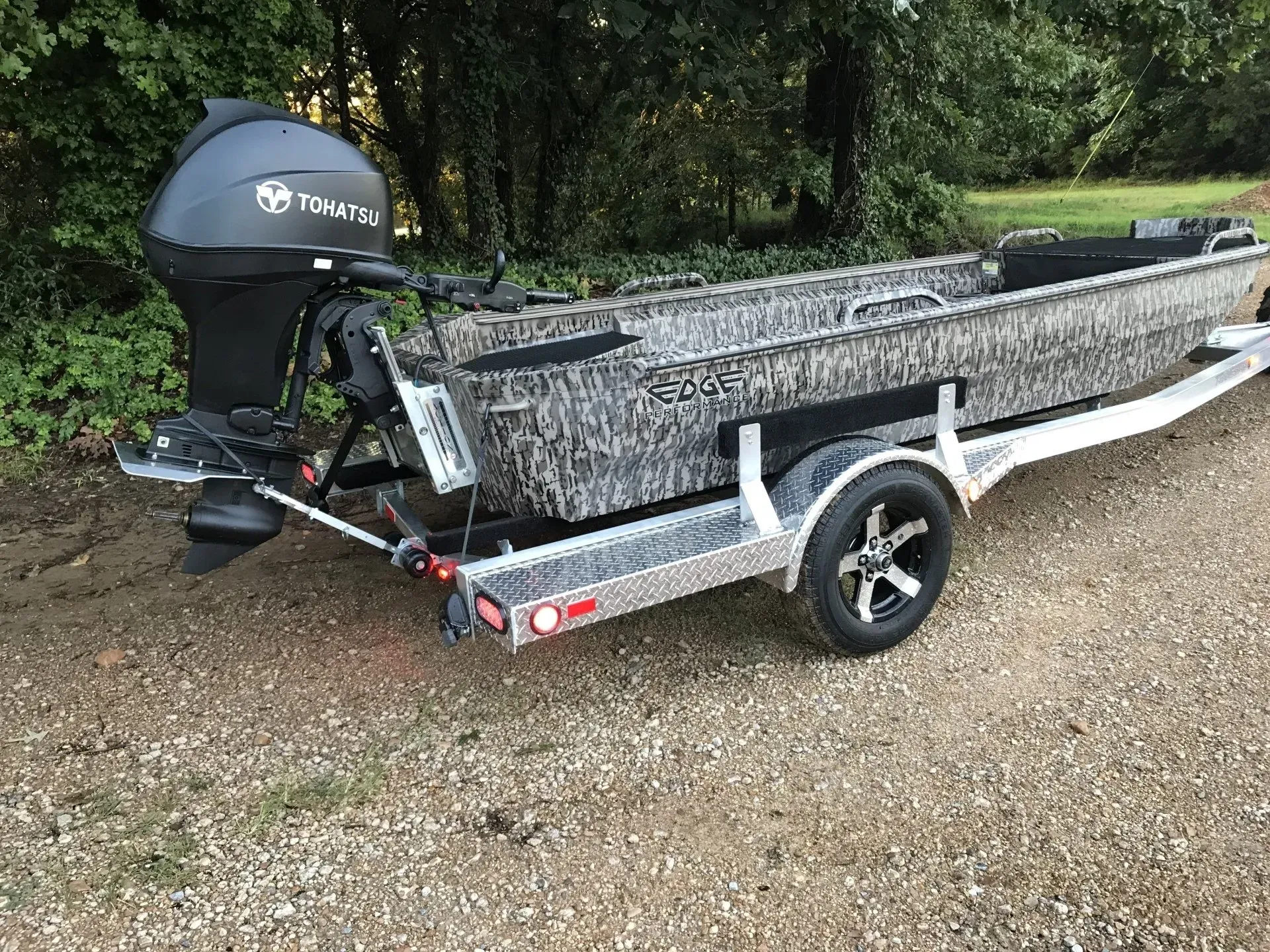 Gray utility boat on a trailer with an outboard motor parked on gravel near trees.