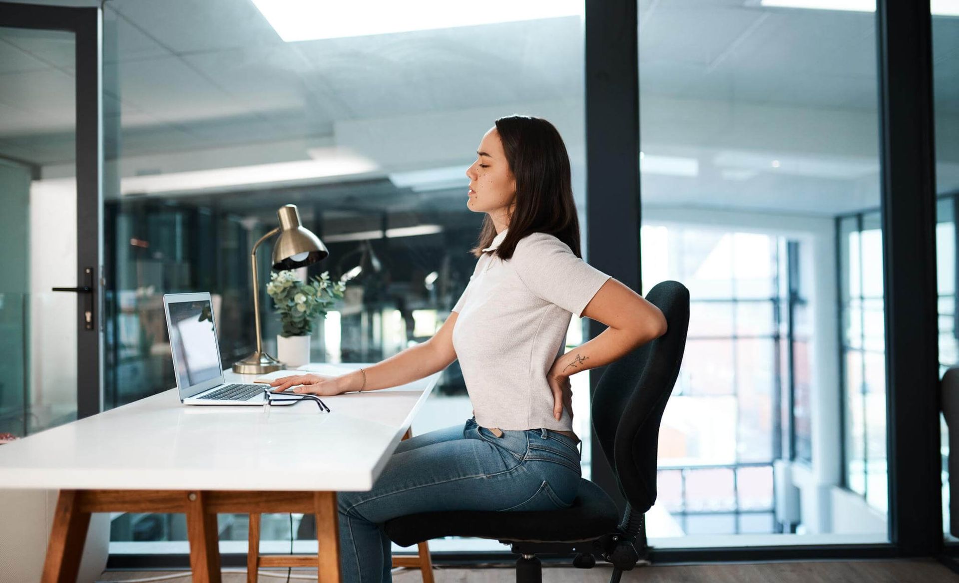 Una mujer está sentada en un escritorio con una computadora portátil y sosteniendo su espalda.