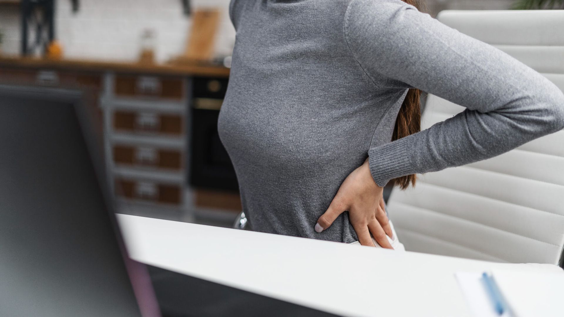 Una mujer está sentada en un escritorio sujetándose la espalda por el dolor.