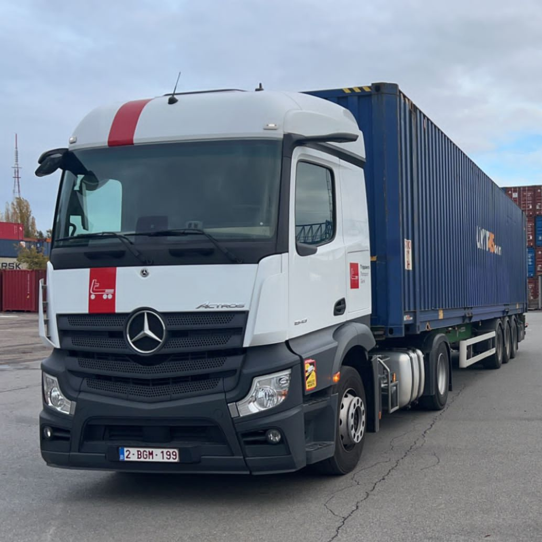 Witte vrachtwagen met oplegger en blauwe container op een verhard terrein; rode strepen op de cabine, bewolkte lucht.